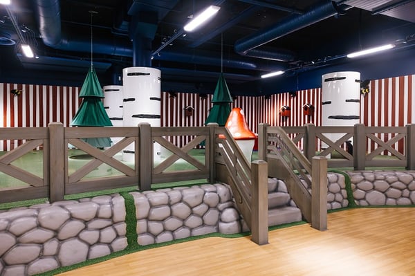 A forest-themed indoor play area with faux stone walls, a wooden fence, and steps leading up to a grassy section featuring white tree-like columns, hanging green tree decorations, and a large red mushroom structure, all set against red-and-white striped walls.