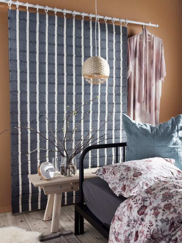 A FOULUM gray and white striped rug and a dressing gown hang on a wall from a curtain rail, behind a bed and a bench.