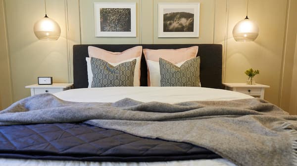 A freshly made bed in the bedroom with various pillows, bedside tables next to it and lighting above.