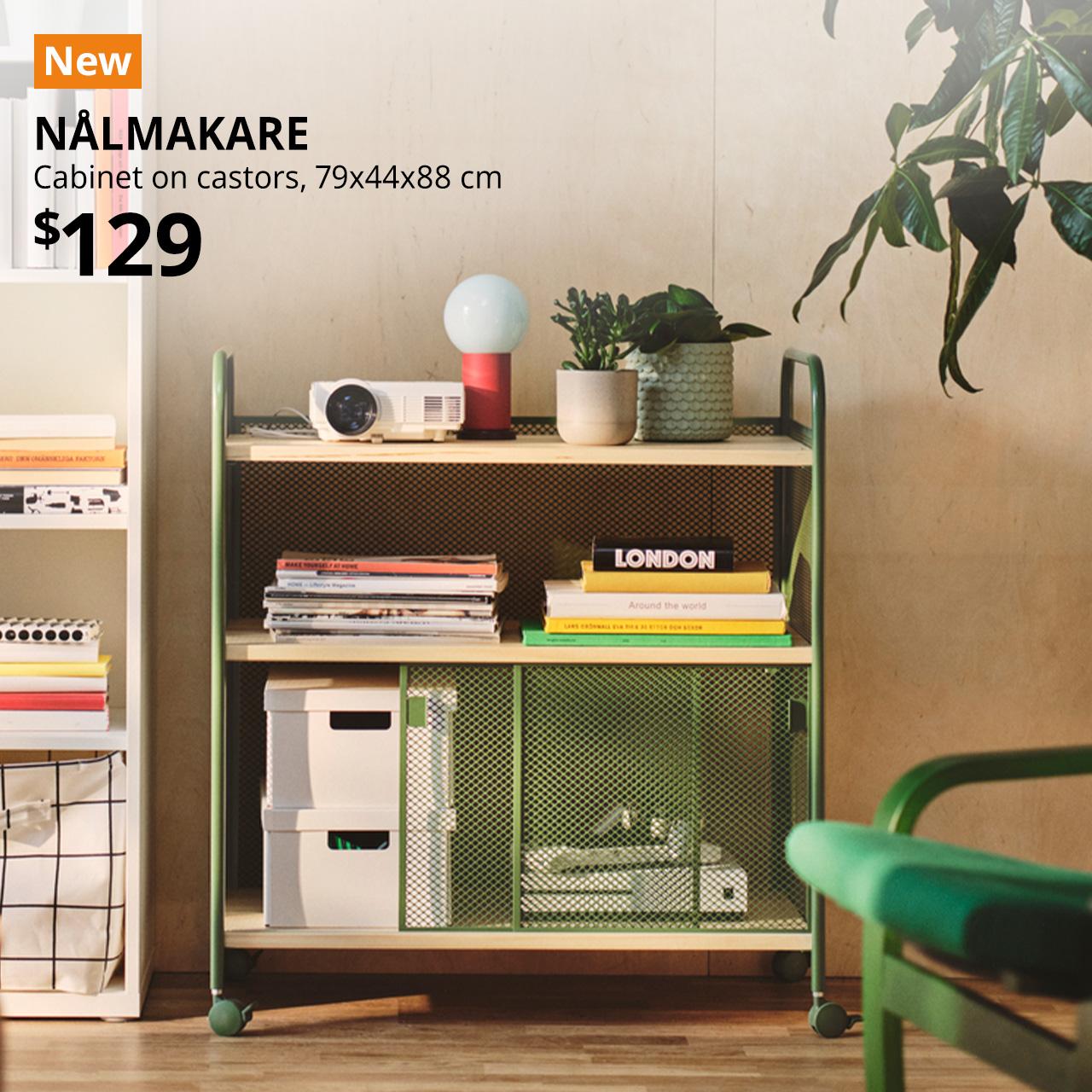 A front-facing view of a dark green NÅLMAKARE cabinet on castors, neatly styled in a living room next to a white BILLY bookcase. The cabinet holds a projector, books, and small storage organisers, showcasing a functional and organised home storage solution. An orange "NEW" label is displayed in the top left corner of the image, featuring white text that reads: "NÅLMAKARE, Cabinet on castors, 79x44x88 cm, $129."