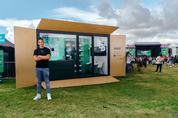 A front view of a life-size flatpack box transformed into a shop called Stay Clean Daily, the owner standing in front of it.
