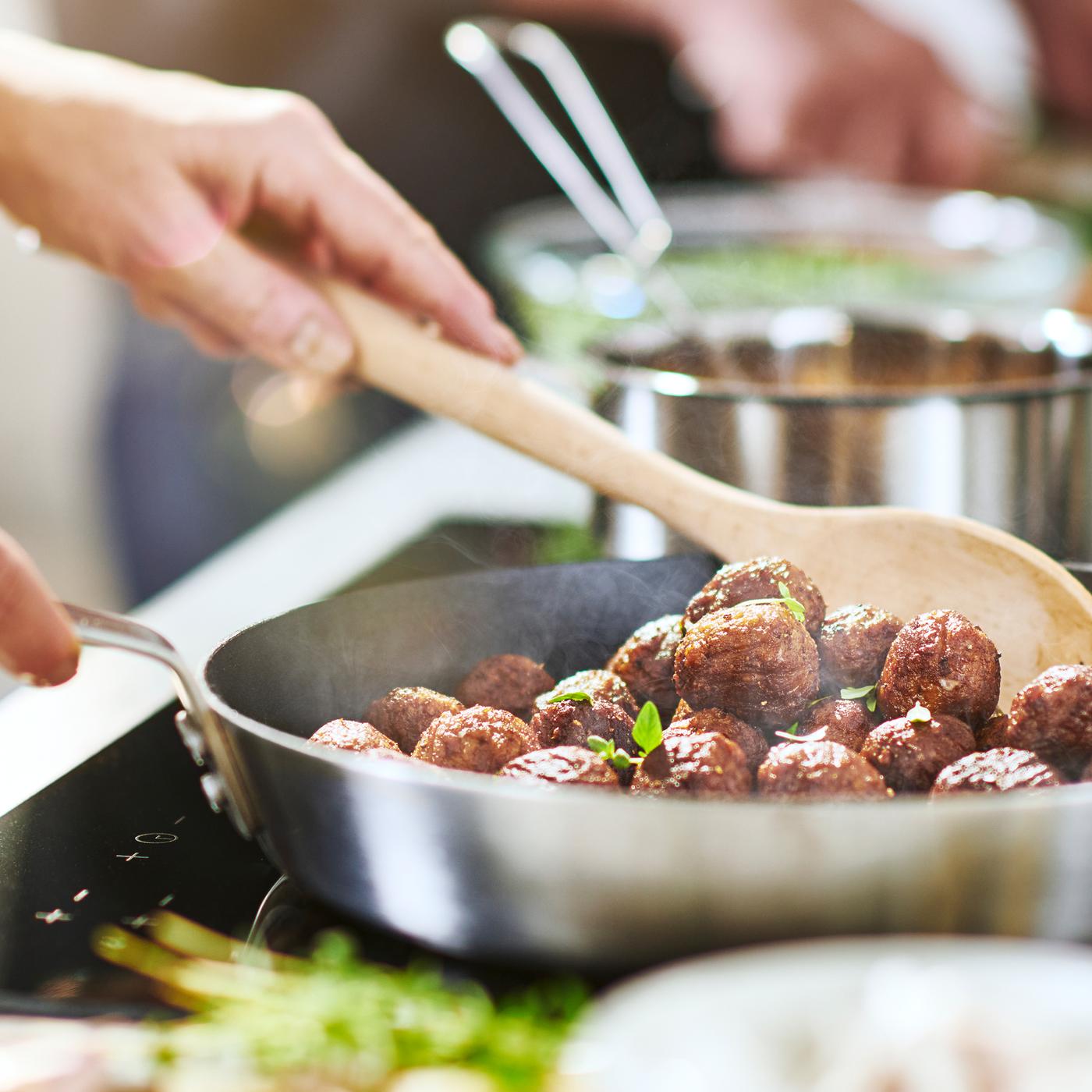 A frying pan full of meatless meatballs cooking on an induction hob whilst being stirred by a person holding a wooden spoon.
