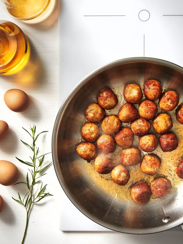 A frying pan of meatballs cooking on a white induction hob. Beside the hob are four eggs, two glasses and a sprig of herbs.