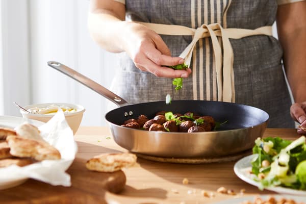 A frying pan with a handle in stainless steel is standing on a hob cooking meatballs in sauce. A wooden spoon is on the side.
