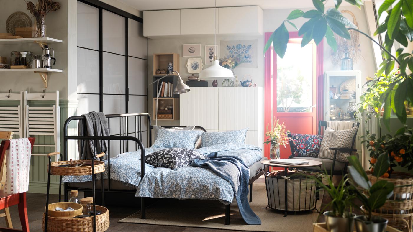 A FYRESDAL day-bed with two mattresses with SOMMARSLÖJA bed linen stands in a small studio apartment near some shelf units.