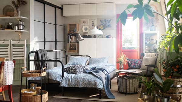 A FYRESDAL day-bed with two mattresses with SOMMARSLÖJA bed linen stands in a small studio apartment near some shelf units.