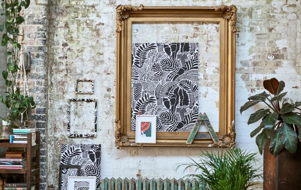 A gallery of framed patterned fabrics displayed on a brick wall with a large gold frame, hanging plants and a low bookcase.