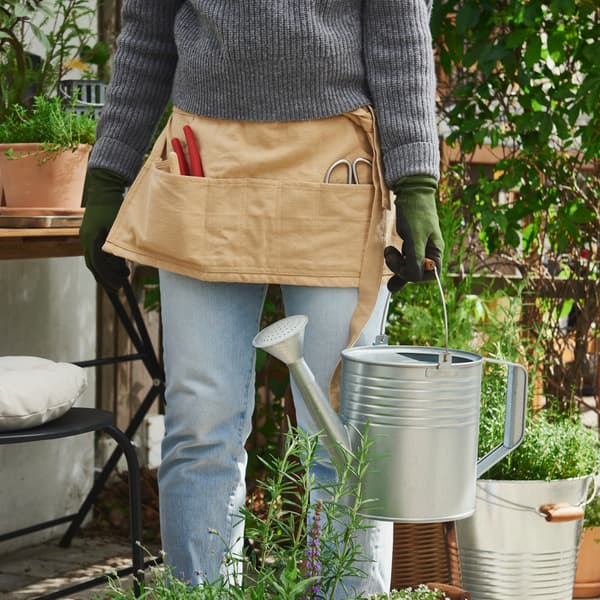 A garden area with plants features a person in a grey sweater holding a KORKGRAN watering can, made of galvanised steel.