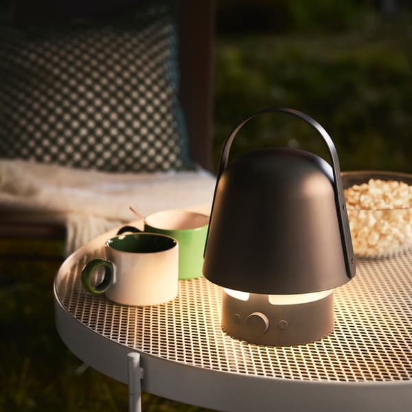 A garden setting features a blue VAPPEBY bluetooth speaker lamp with a rounded design, standing on a table with drinks.