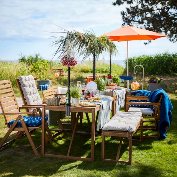 A garden setting with a light brown wooden NÄMMARÖ table with food and drinks on a sunny day. Chairs, a bench and a parasol.
