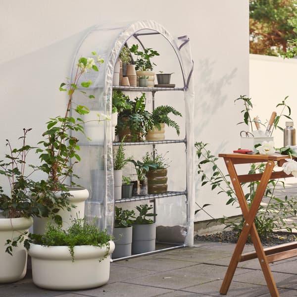 A garden with a transparent white ASPAREN greenhouse filled with plants on shelves next to a wooden table and green plants.