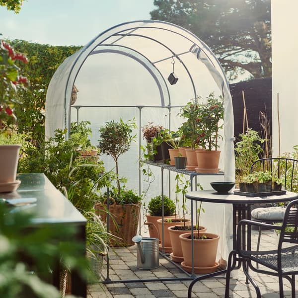 A garden with an ASPAREN greenhouse in transparent white, surrounded by potted plants and outdoor seating.