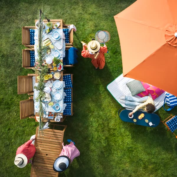 A garden with outdoor seating features a light brown stained NÄMMARÖ table surrounded by people enjoying refreshments.