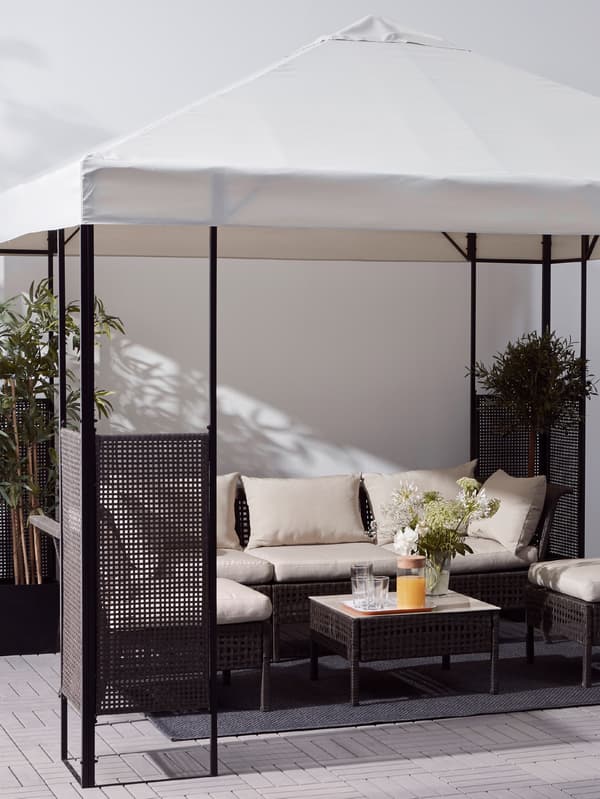 A gazebo with a white roof and dark brown frame, an outdoor sofa and chairs with cushions, and a table with plants.