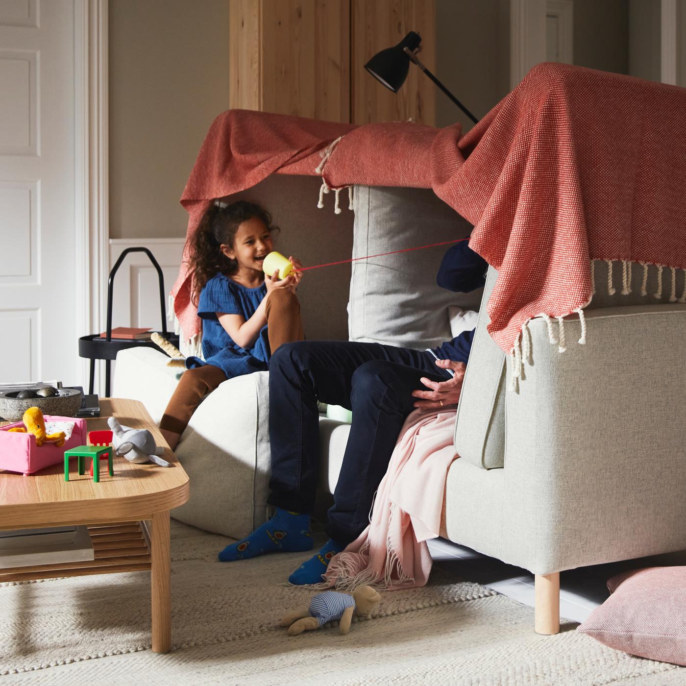 A girl and a man playing inside a blanket fort built on a beige PÄRUP sofa, beside an oak veneer coffee table on a beige rug.