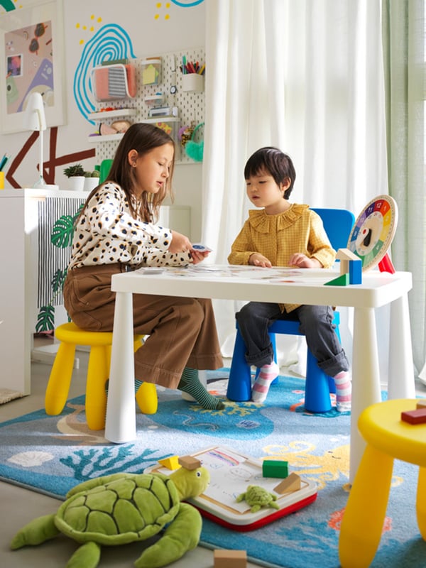 A girl and her younger brother sit on MAMMUT stools around the MAMMUT white children's table. She teaches him how to identify time using the UNDERHÅLLA educational wooden clock. A BLÅVINGAD rug with patterns of ocean animals is placed on the floor.