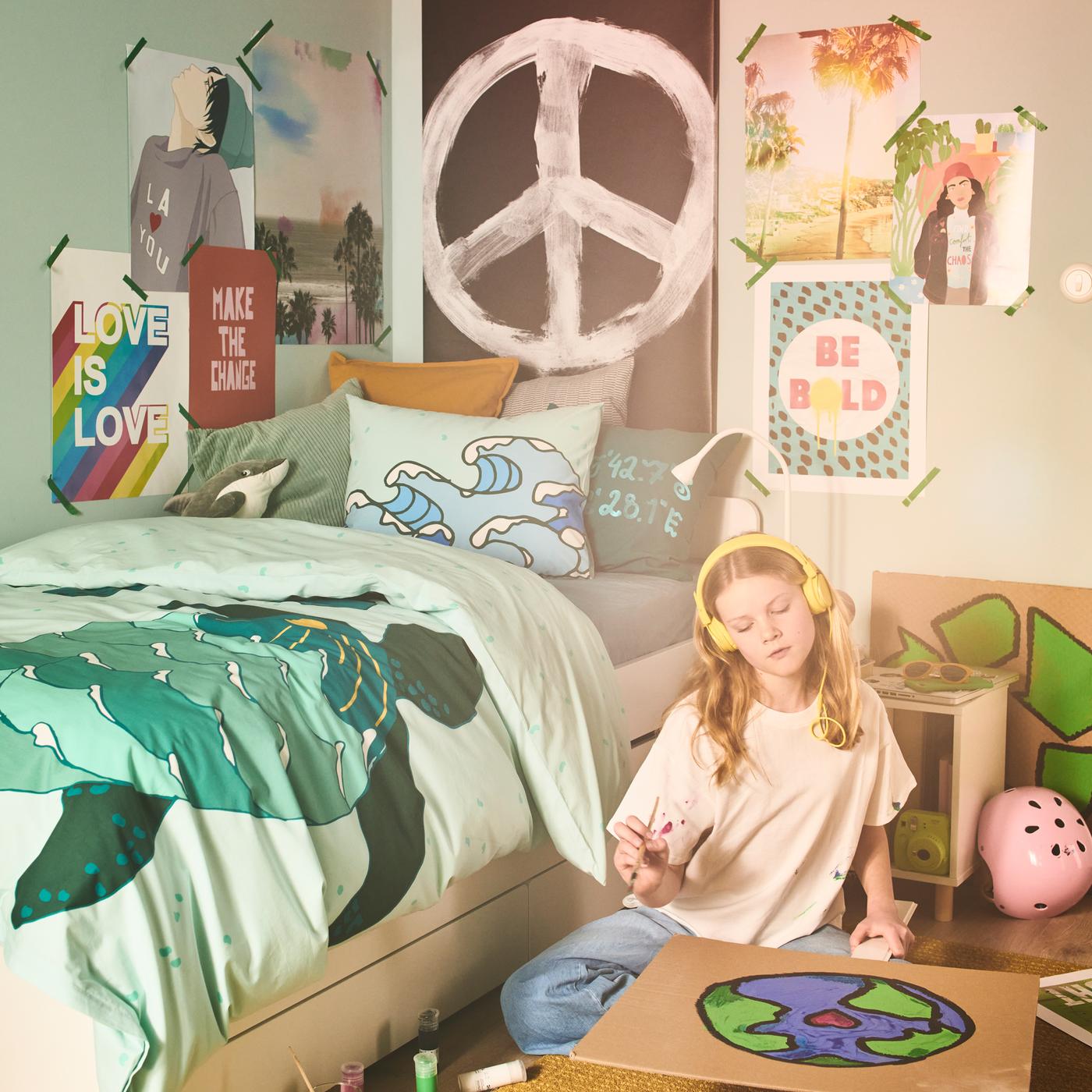 A girl, colouring in a poster of the world, sits on the floor beside a bed dressed in a BLÅVINGAD turtle duvet cover.