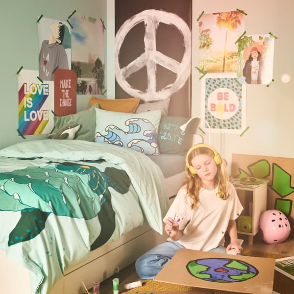 A girl, colouring in a poster of the world, sits on the floor beside a bed dressed in a BLÅVINGAD turtle duvet cover.