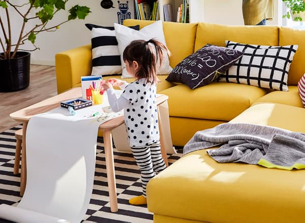 A girl drawing won a long roll of paper on top of a coffee table and next to a yellow IKEA VIMLE three-seat sofa sectional.