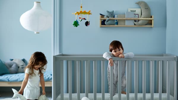 A girl holding two soft toys beside a toddler standing in a grey crib, with a children’s bed with blue bed linen behind.