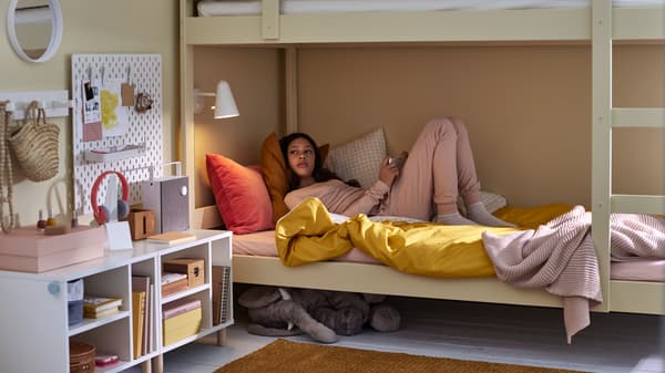 A girl in a bottom bunk bed with bed linen in mixed colors, a soft toy elephant under the bed and various white storage.