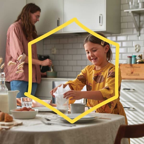 A girl is standing at a table folding napkins, and behind her is an adult woman standing in the kitchen stirring a bowl with a spoon.