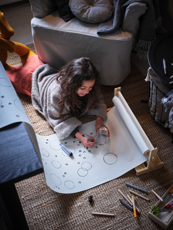 A girl lies on the floor, drawing shapes on a long roll of white paper to make a tablecloth for a festive dinner.