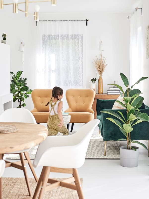 A girl runs through an open-plan dining/living area with green velvet sofa, mustard easy chairs and white dining chairs.