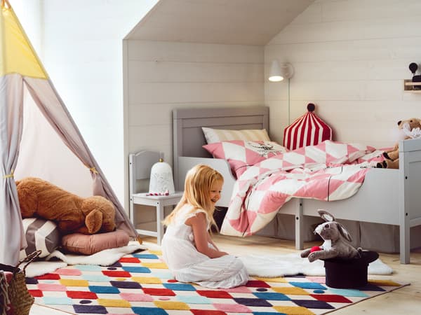 A girl sits on a brightly colored BUSENKEL rug in a bedroom near a grey SUNDVIK extendable bed and looks at a toy animal.