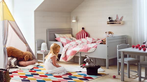A girl sitting on a brightly coloured rug in a bedroom containing a grey SUNDVIK extendable bed and a HÖVLIG play tent.