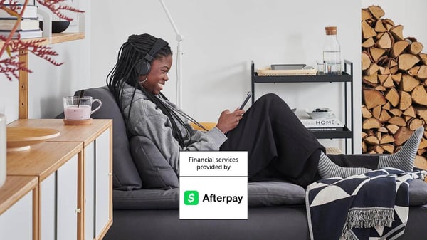 A girl sitting on a chaise with a mug next to her and smiling at her phone with headphones on.