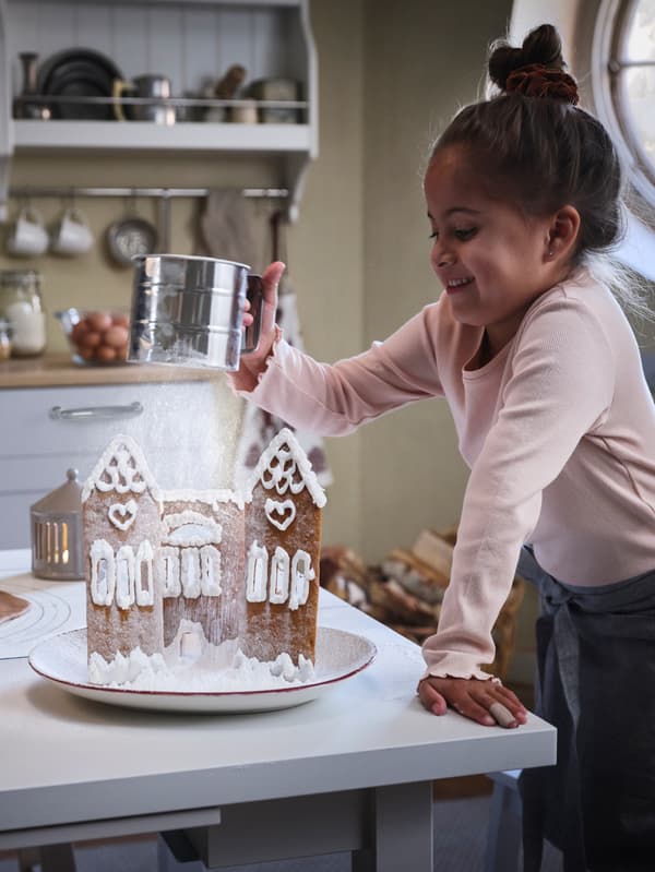 A girl uses an IDEALISK flour sifter to sprinkle sugar like a snowstorm on top of an iced gingerbread house.