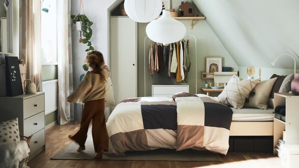 A girl walking into a bedroom with a bed made with brown/grey checkered bed linen, plus various storage and white pendants.