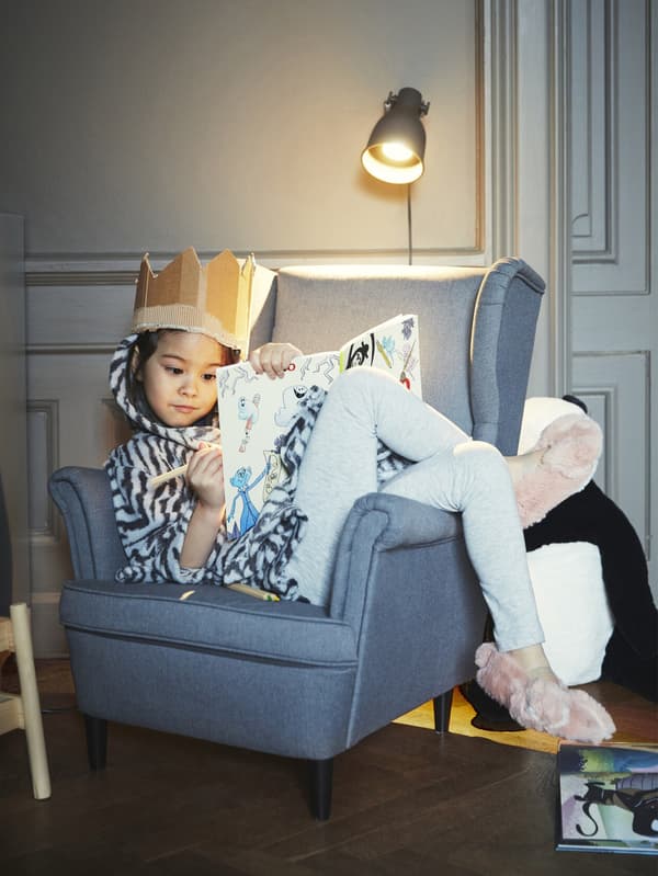A girl wearing a cardboard crown, colouring while curled up on a grey chair.