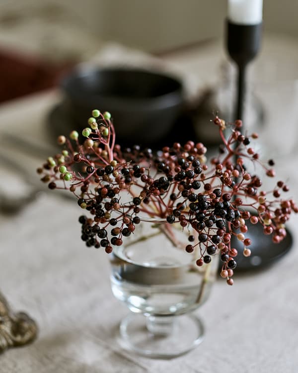A goblet style glass used as a vase with a bouquet of wild berries, on a table covered in linen with candles and crockery.