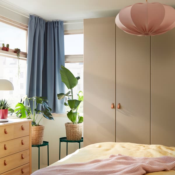 A gray-beige wardrobe, blue curtains, a pink pendant lamp, yellow bed textiles, a pale pink throw and two green tray tables.