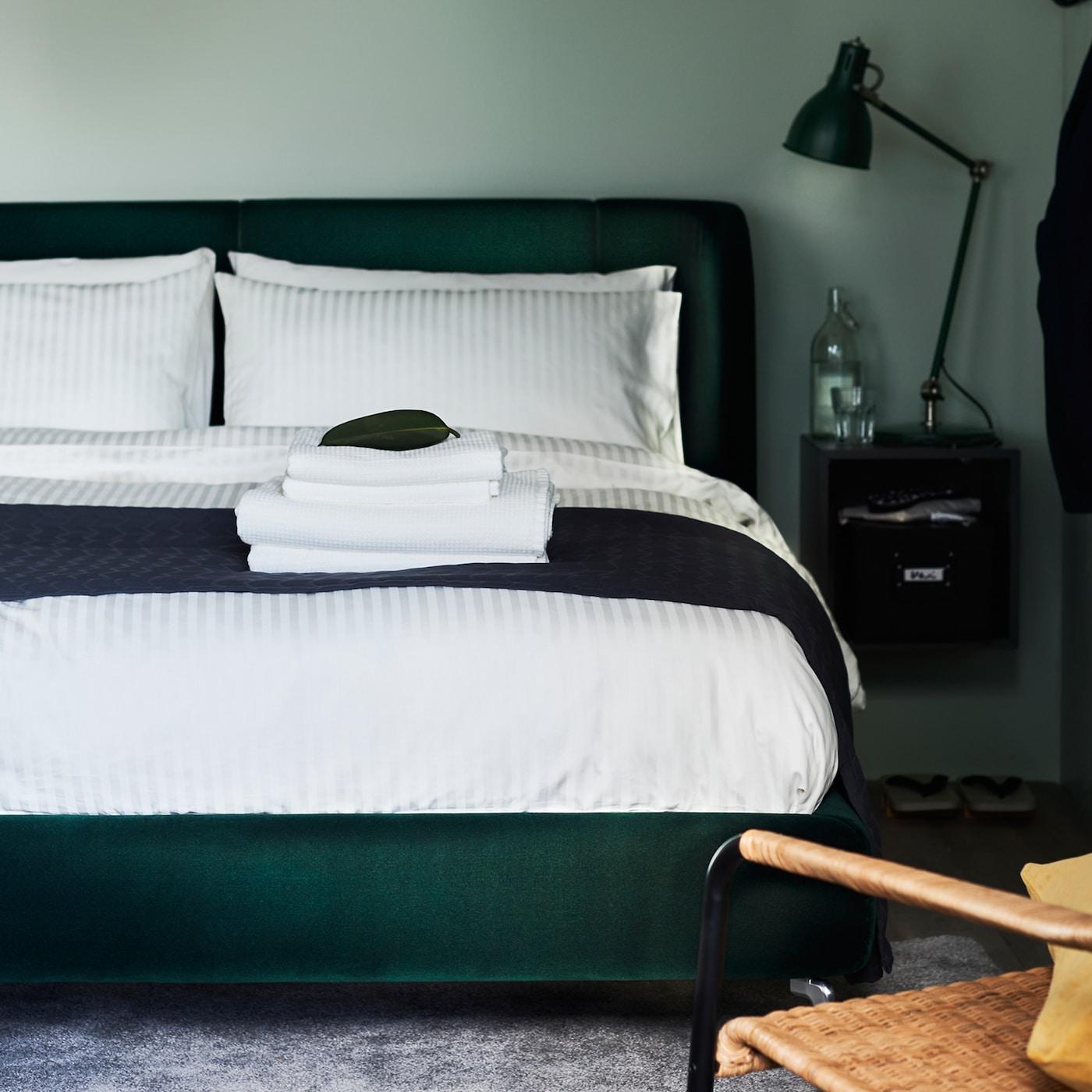 A green bedroom with a dark green upholstered bed covered in white bed linen. A black work lamp is on a bedside table.