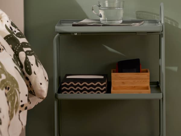 A green bedside table with a glass of water on top and organisation accessories on the shelf under.
