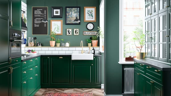 A green kitchen with a white sink with a visible front, a brass-coloured tap, a gas hob, and two black ceiling lamps.