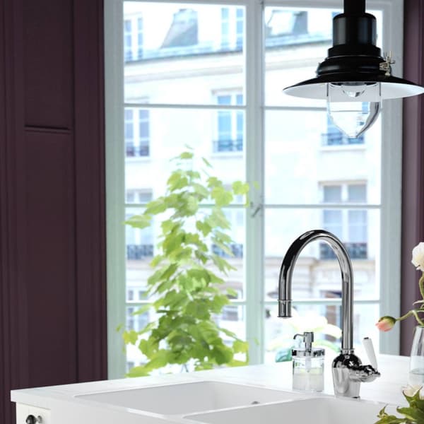 A green kitchen with white tiles, a brass-coloured mixer tap, a white ceramic sink and an ivory/gold-coloured watering can.