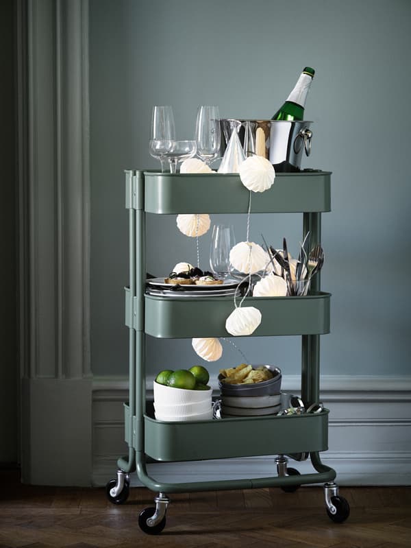 A green RÅSKOG trolley is packed with things for a party, including glasses, dinnerware and drinks to share.