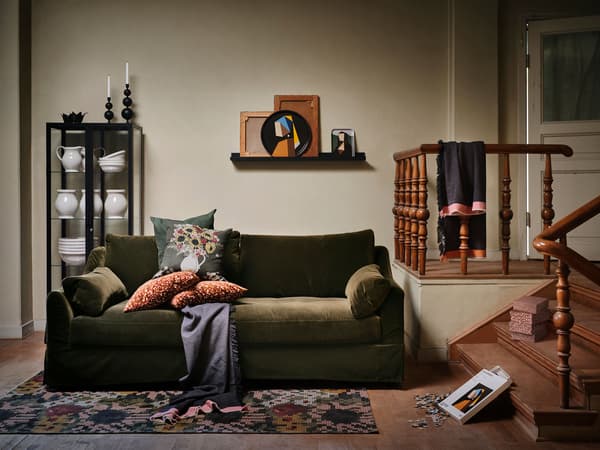 A green sofa in the middle of a room with hardword floors and stairs to the right, and glass storage cabinets behind it.
