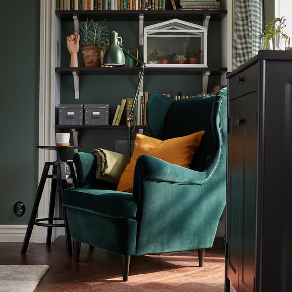 A green STRANDMON wing chair stands by shelves with books, a green floor lamp, a black cabinet and a black barstool.