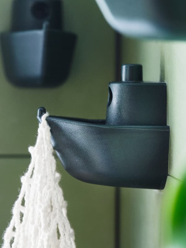 A green tile wall with three green KNOPARE hooks resembling a toy boat, one of which is holding a white macrame bag.