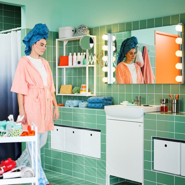 A green-tiled bathroom with a white BJÖRKÅN sink and NYSJÖN wash basin cabinet with one door and a woman wearing pyjamas.