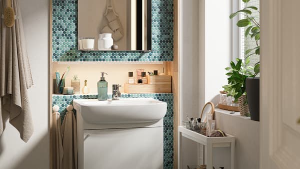A green-tiled bathroom with a white wash-basin cabinet, mirror with shelf and white trolley with toiletries in it.