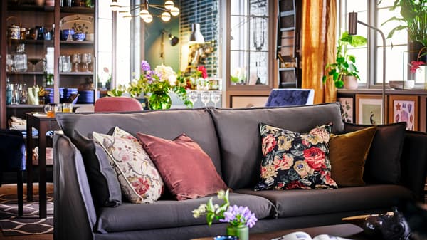 A grey BACKSÄLEN sofa with four scatter cushions in front of a dining table and chairs with framed art on the wall.