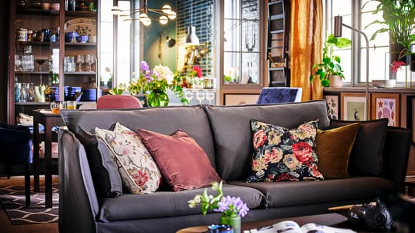 A grey BACKSÄLEN sofa with four scatter cushions in front of a dining table and chairs with framed art on the wall.