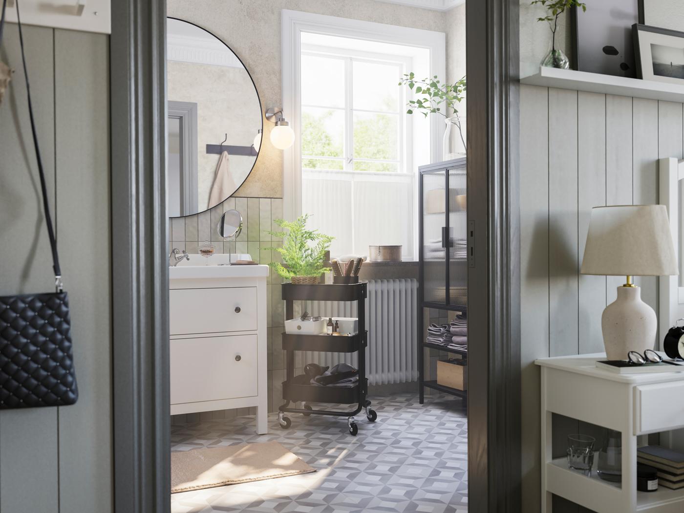 A grey bathroom with a big window has a white HEMNES vanity, a black RÅSKOG cart and a MOSSJÖN glass door cabinet.