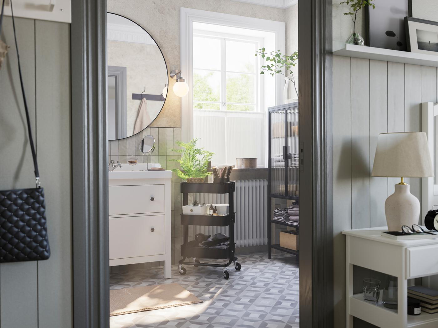 A grey bathroom with a big window has a white HEMNES washstand, a black RÅSKOG trolley and a MOSSJÖN glass door cabinet.